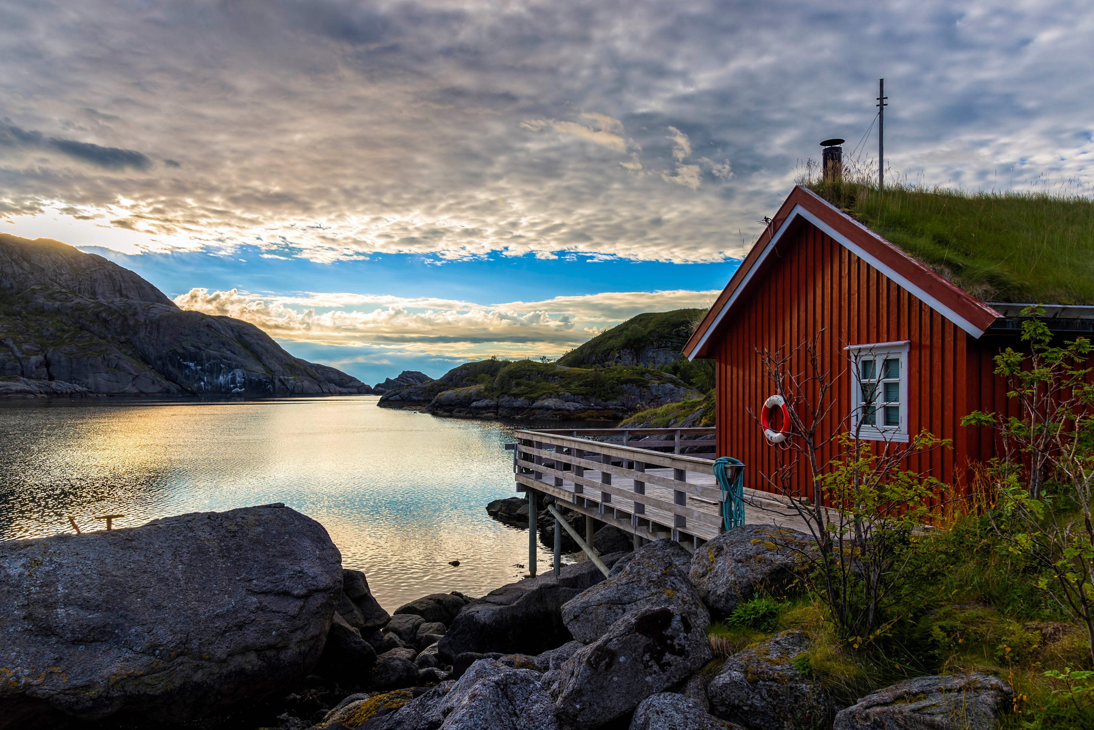Rotes Haus mit Grasdach am rechten Rand des Bildes mit weißen Fenstern und einem Rettungsring an der Wand. Das Haus sieht sehr alt aus. Es gibt eine Veranda vor dem Haus, die über einem Fjord gebaut ist. Vor dieser Veranda sieht man auf einen Fjord, der nach hinten ins Meer mündet. Im Vordergrund gibt es große Felsen, die sich nach links über das Bild verteilen. Am Himmel gibt es kleine weiße Wolken.